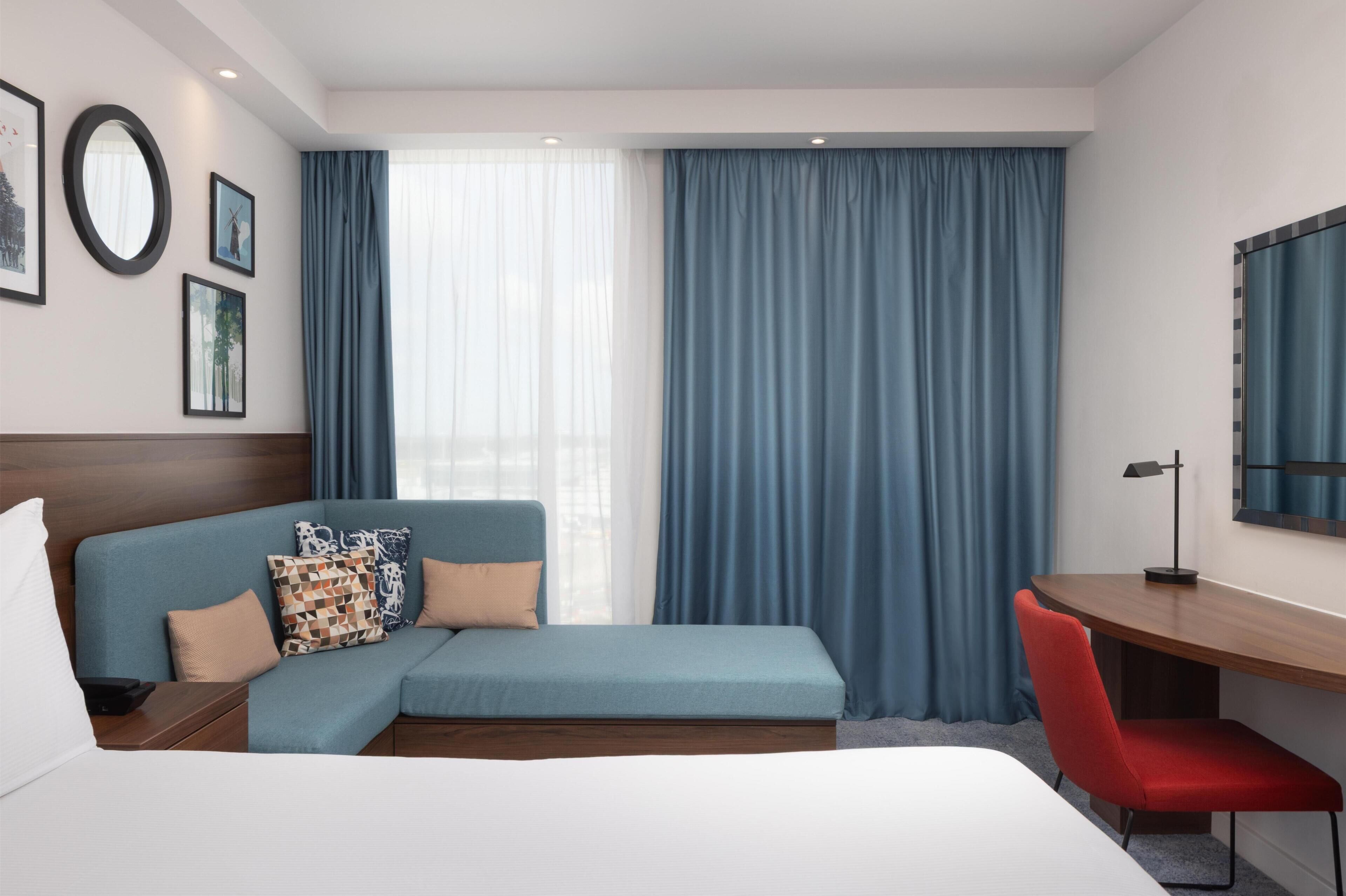 A modern hotel room with a bed, a sofa, a desk, and a TV.