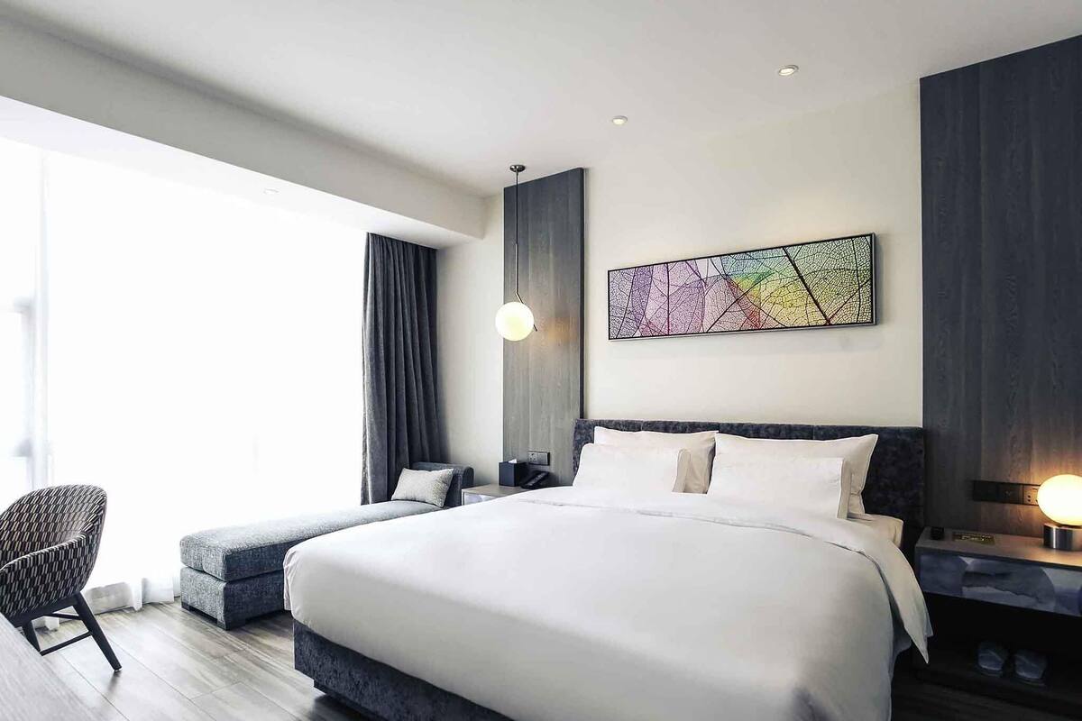 A modern hotel room with a large bed, a seating area with a chair, a bedside table with a lamp, and a large window.
