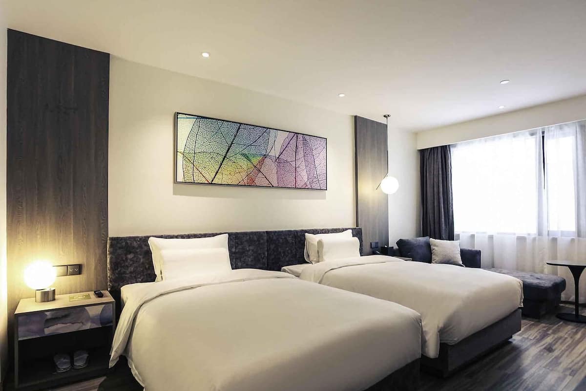 A hotel room with two beds, a large abstract painting on the wall, a bedside table with a lamp, and a view of the outdoors through the window.