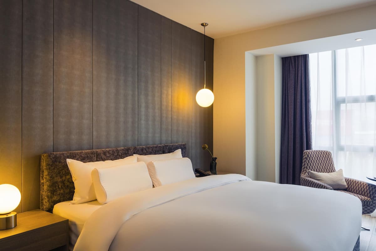 A hotel room with a large bed, a nightstand, a lamp, a chair, and a window with curtains.