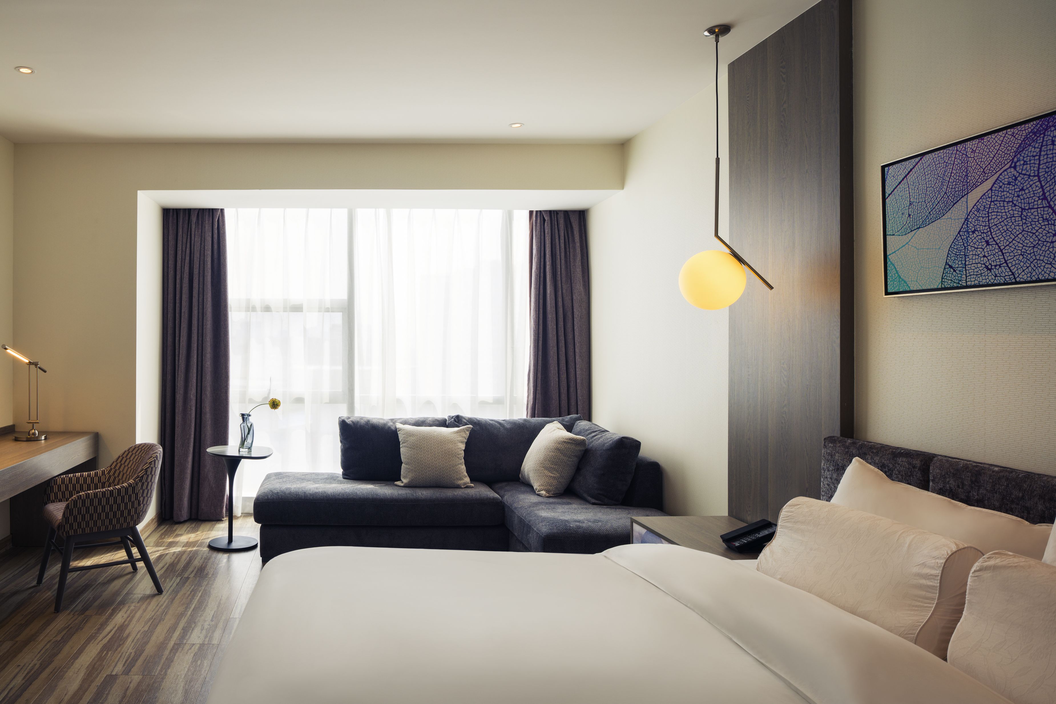 Een moderne hotelkamer met een groot bed, een bank, een bureau en een stoel. Er is een raam met gordijnen, een lamp en een schilderij aan de muur.