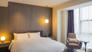 Ein modernes Hotelzimmer mit einem großen Bett, einem Sessel, einem kleinen Tisch und Blick auf die Stadt durch große Fenster.