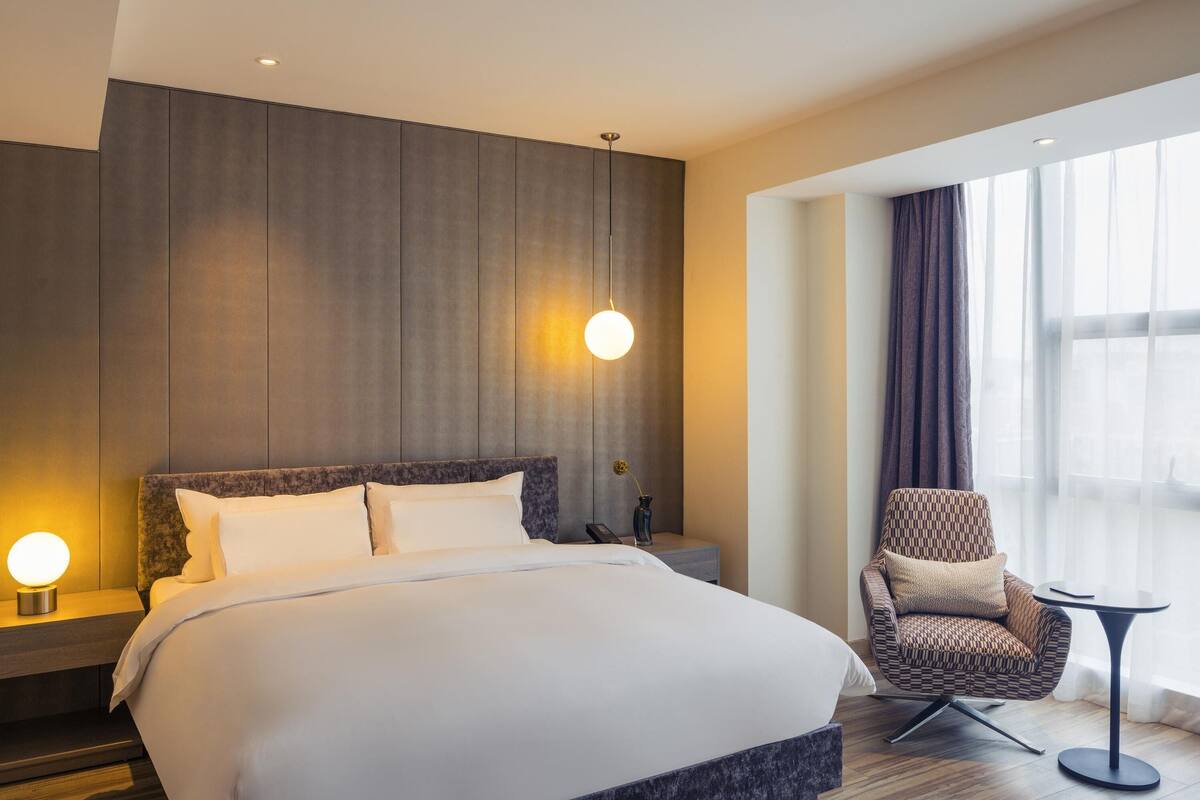 A modern hotel room with a large bed, a chair, a small table, and a view of the city through large windows.
