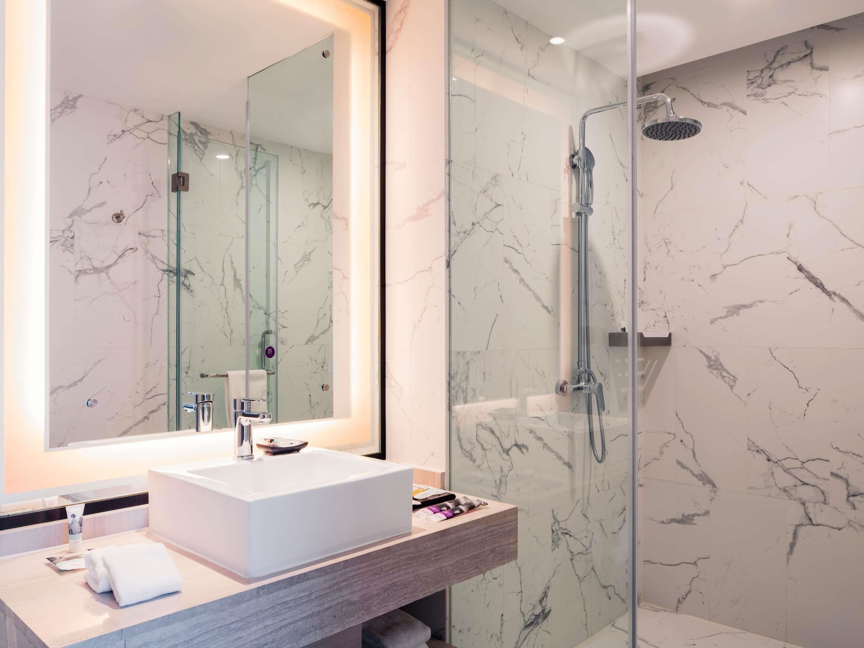 Un baño moderno con un espejo grande, un lavamanos blanco, ducha con mampara de vidrio y paredes de mármol.