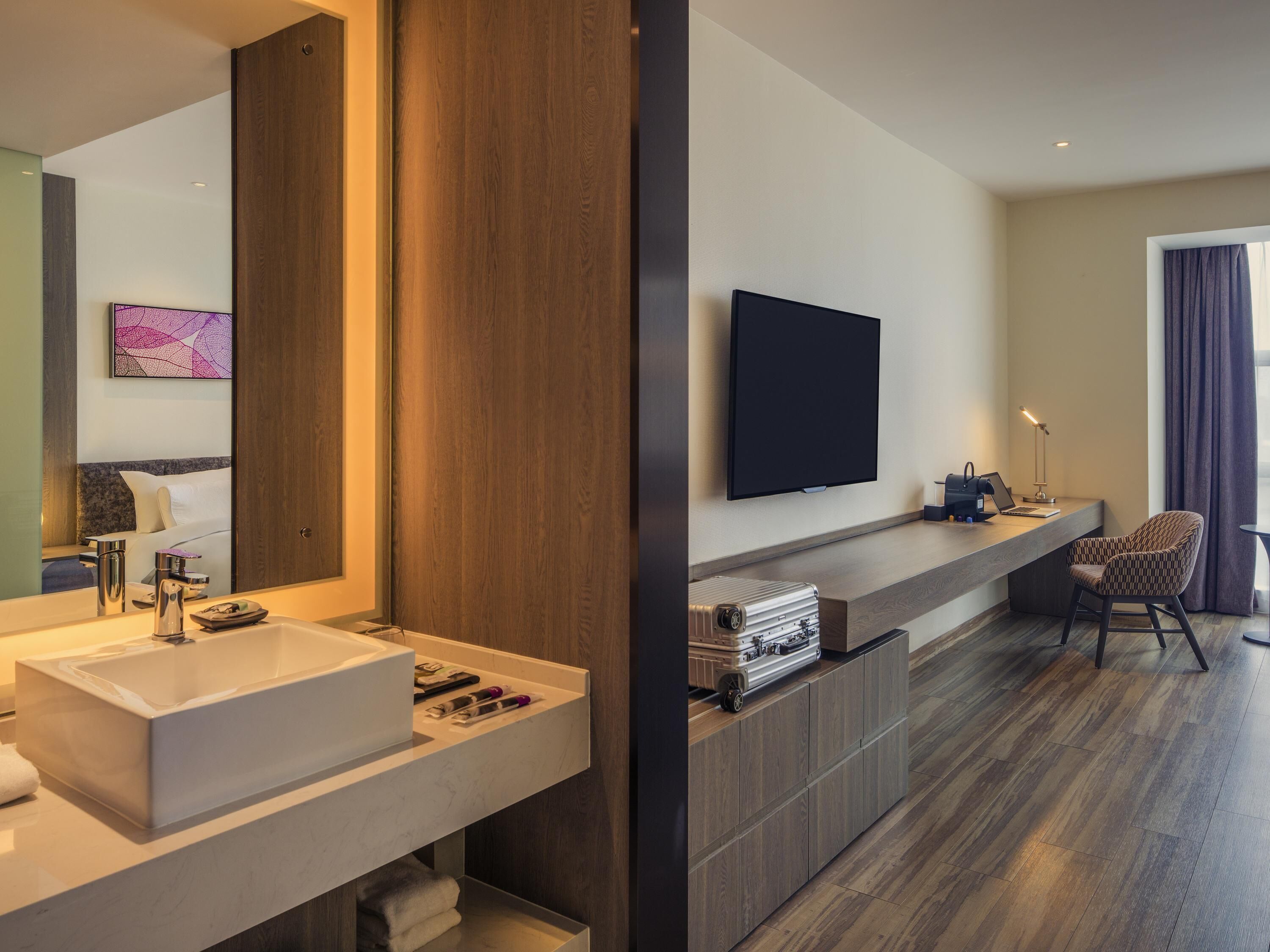 A modern hotel room with a large mirror, a flat-screen TV, a desk with a laptop, a chair, and a wooden cabinet.