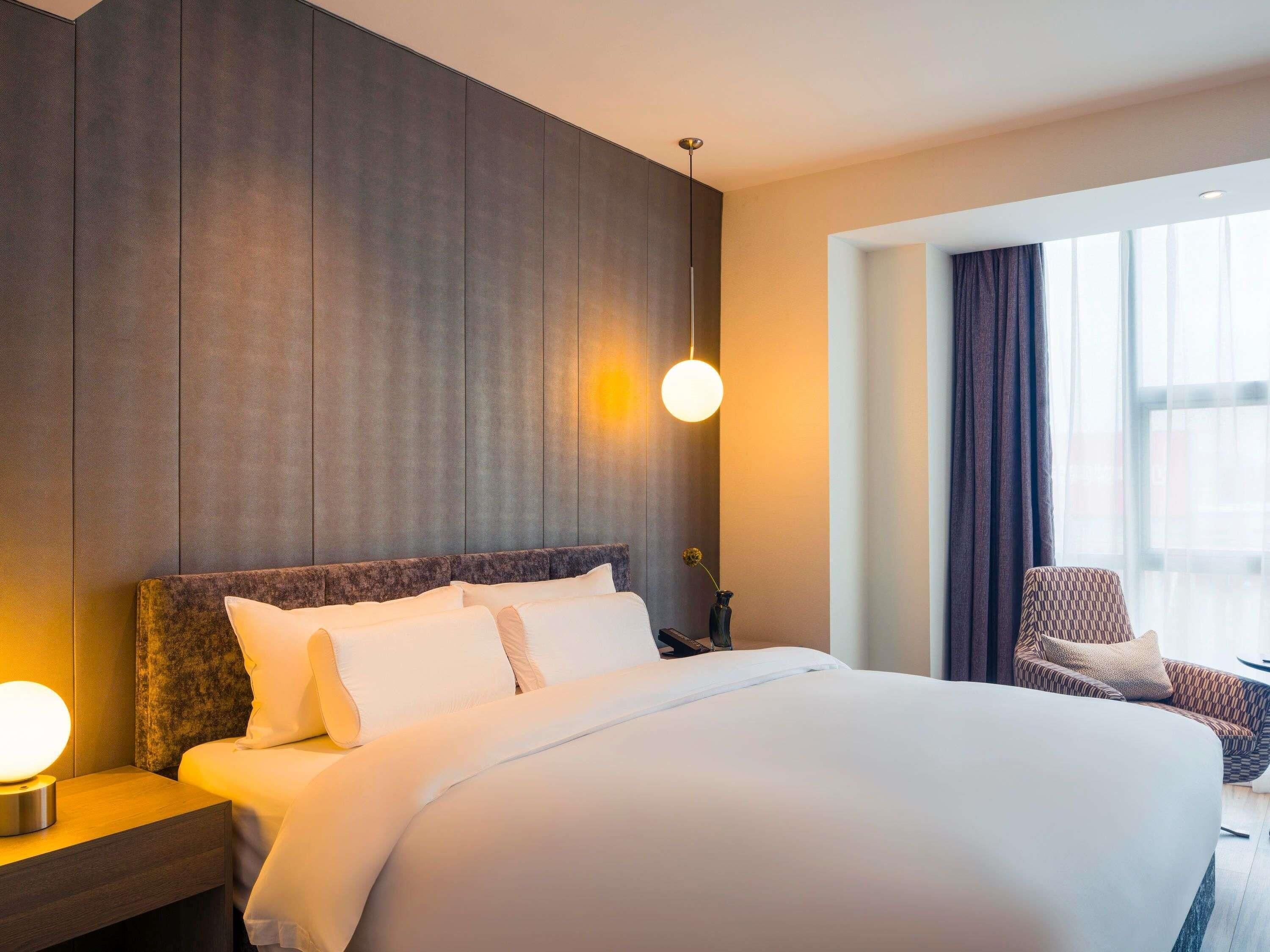 Une chambre d’hôtel moderne avec un grand lit, une table de chevet avec une lampe, une chaise et une fenêtre avec des rideaux.