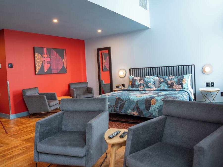 A modern hotel room with a red accent wall, a large bed with patterned bedding, and two grey armchairs.