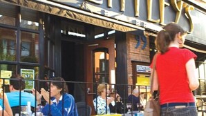 Un espace extérieur aménagé avec des tables et des chaises, où les clients d’un restaurant peuvent s’asseoir, et une personne qui passe.