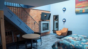 Une pièce moderne avec un mur en briques, un escalier, un téléviseur à écran plat, une table ronde, deux chaises, un canapé et un tapis coloré.