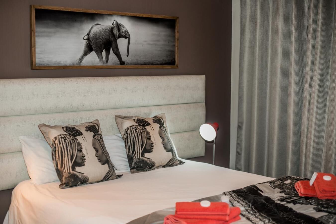 Habitación de hotel con una cama grande, dos almohadas con un diseño de retrato, cabecera, un cuadro enmarcado de un elefante, una lámpara roja y una ventana con cortinas.