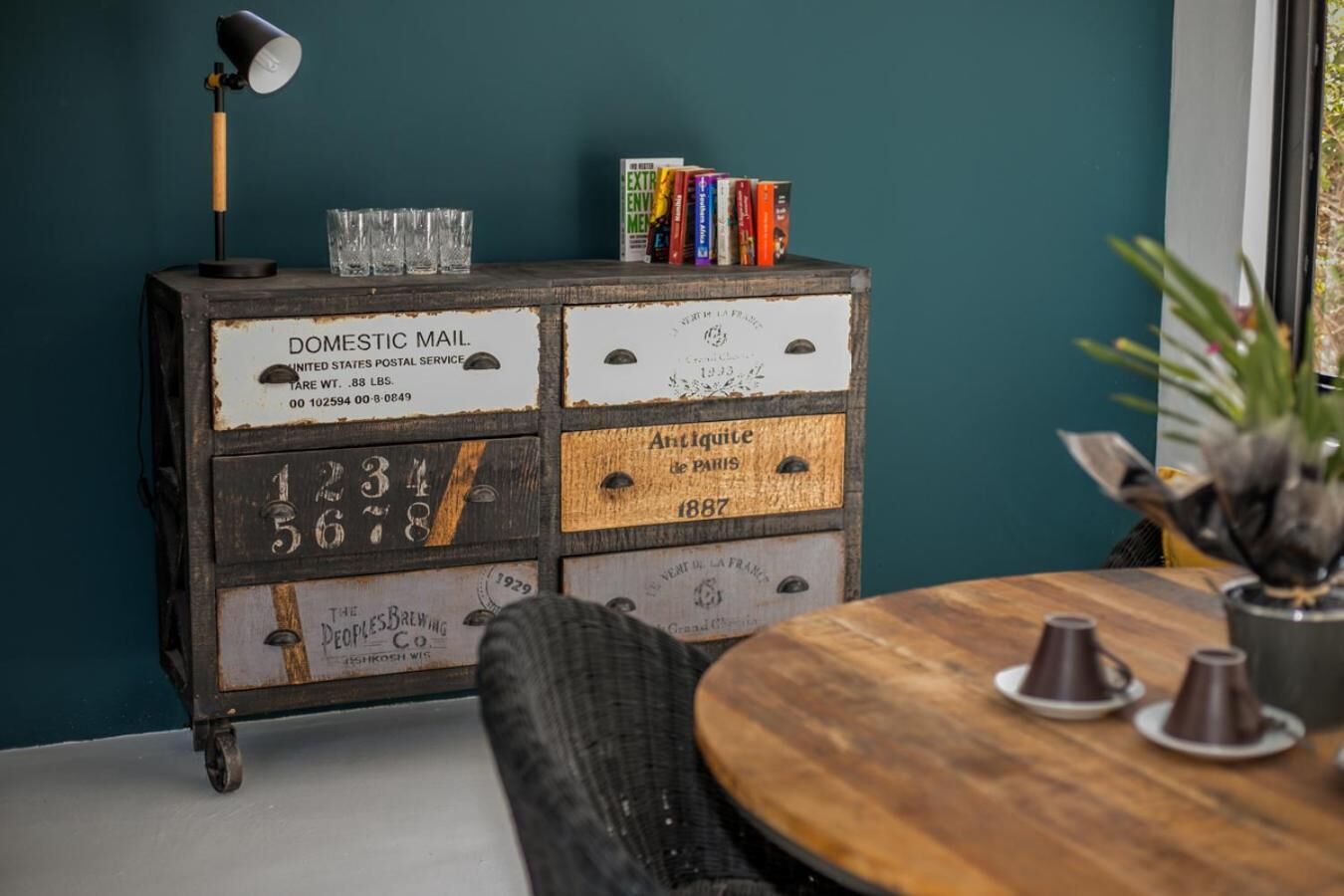 Une pièce comprenant un vieux buffet en bois où sont exposées diverses étiquettes et des livres, une lampe et une table ronde en bois sur laquelle est posée une plante en pot.