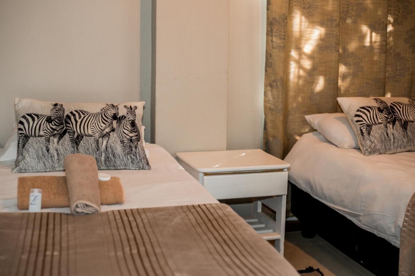 Habitación de hotel con dos camas, almohadas con estampado de cebra, una mesita de noche pequeña y blanca, y una toalla enrollada sobre la cama.