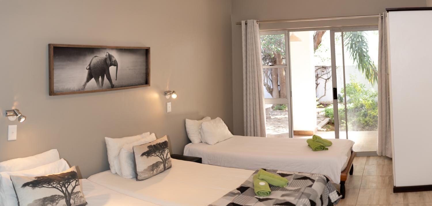 A hotel room with two beds, a large window with curtains, a painting of an elephant, and a view of greenery outside.