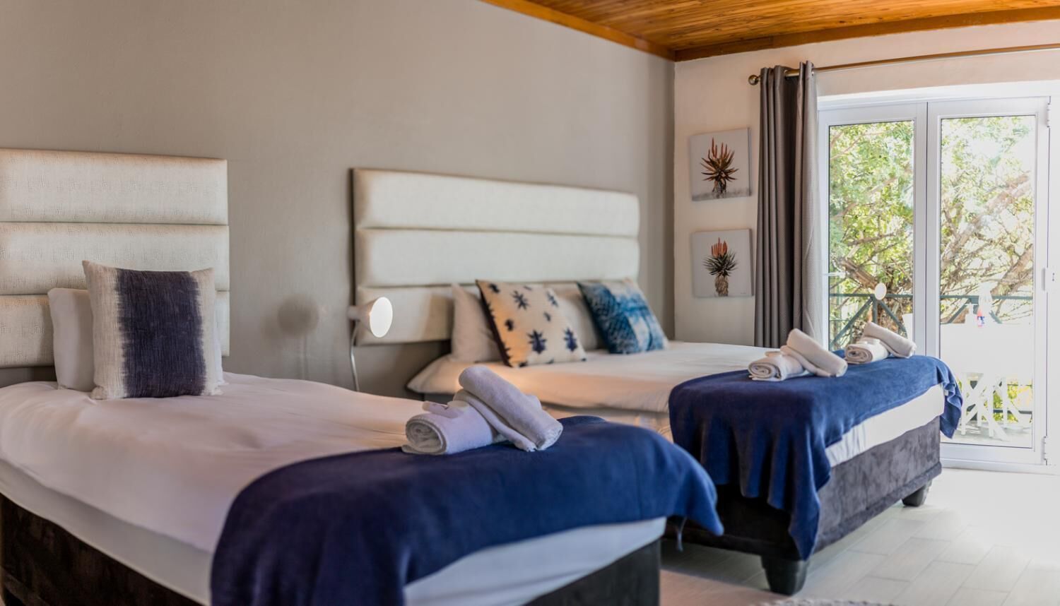 A hotel room with two beds, a large window with curtains, and a view of trees outside.