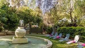 Un jardín con una fuente, sillones de descanso y un edificio al fondo.