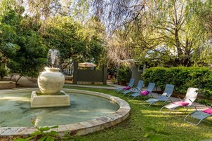 Un jardín con una fuente, sillones de descanso y un edificio al fondo.