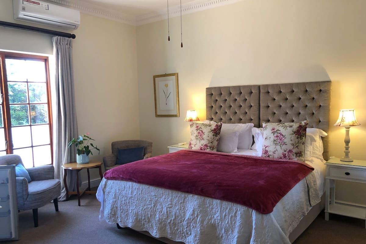 Een hotelkamer met een groot bed, een stoel, een klein tafeltje, een nachtkastje en een raam met gordijnen.