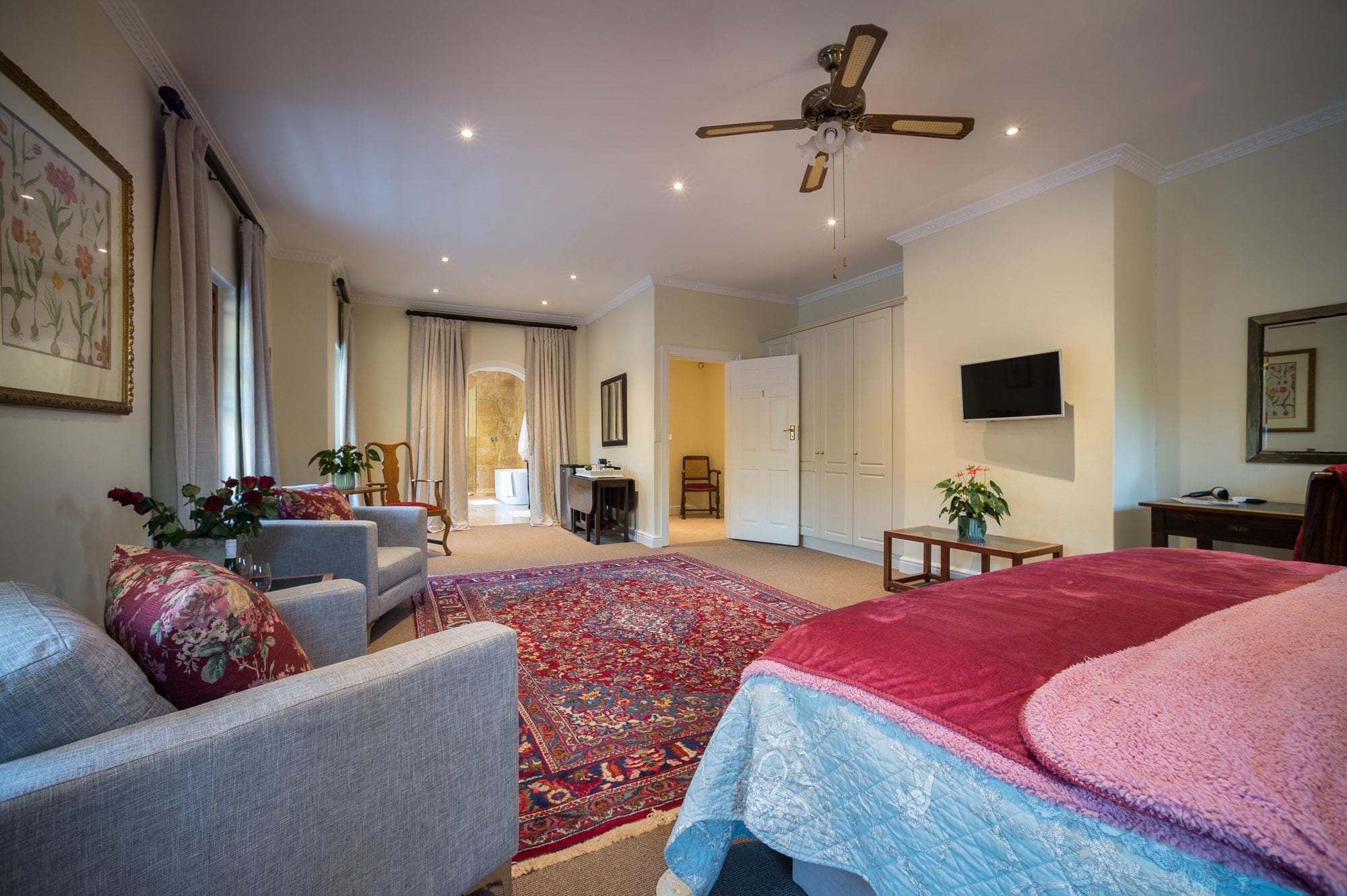 A spacious hotel room with a large bed, a sitting area with a chair and a small table, a television mounted on the wall, and a ceiling fan.