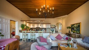 Een ruime woonkamer met een houten plafond, een eethoek met geruit tafelkleed, en een keuken met witte kasten en een houten kookeiland.