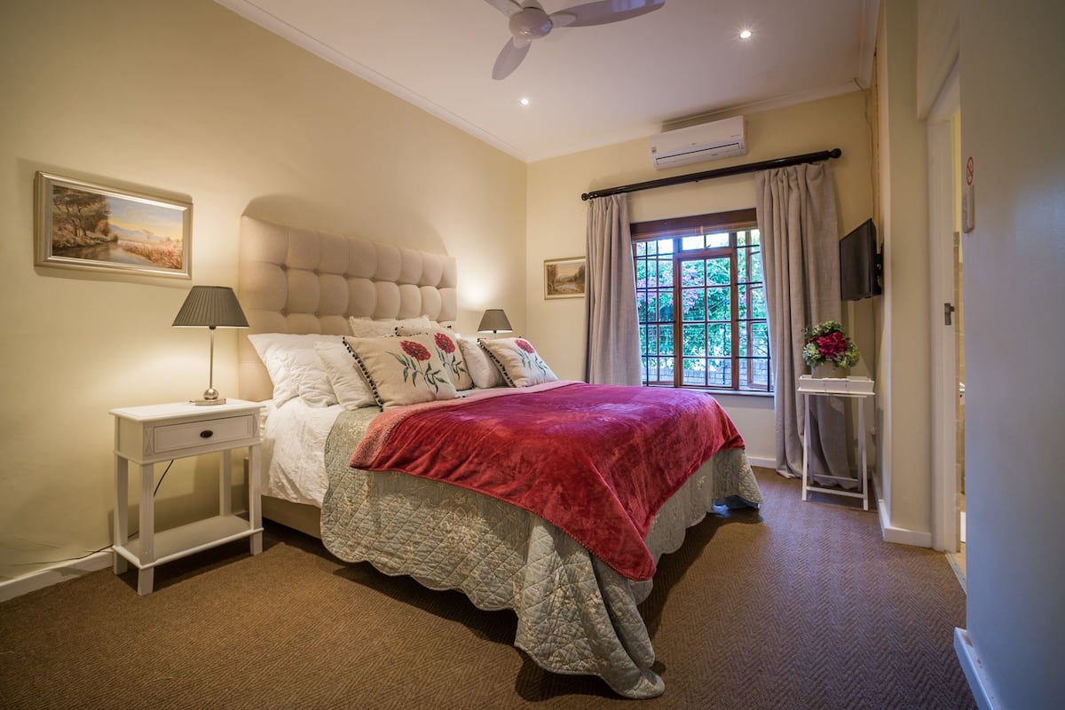 Een slaapkamer met een groot bed, een nachtkastje, een plafondventilator, een airconditioning en een raam met gordijnen, plus een schilderij aan de muur.