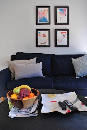 A living room with a dark blue sofa, a wooden fruit bowl, and a remote control on a magazine.