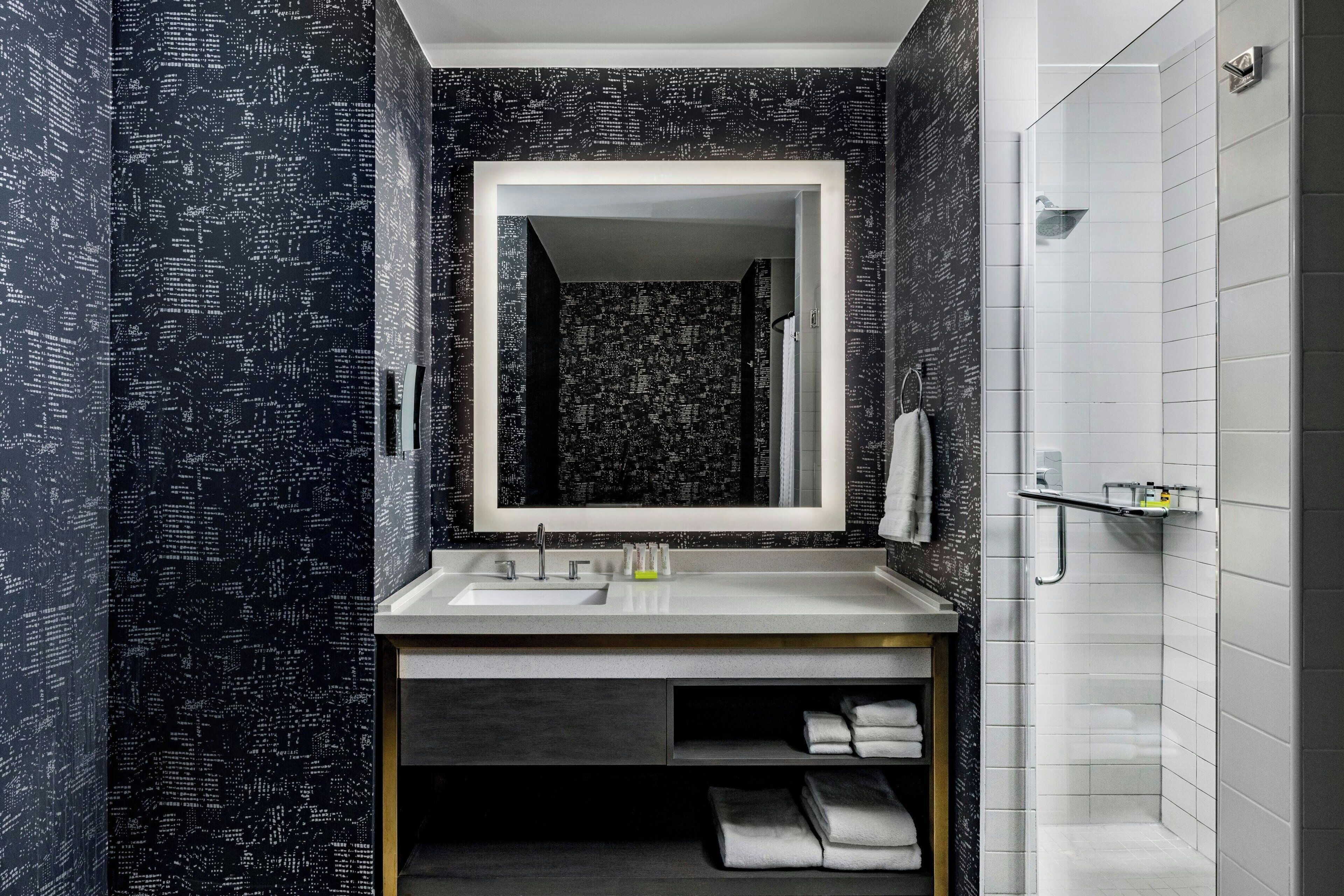 A modern bathroom with a dark patterned wallpaper, a large mirror, a white sink, and a shower area.