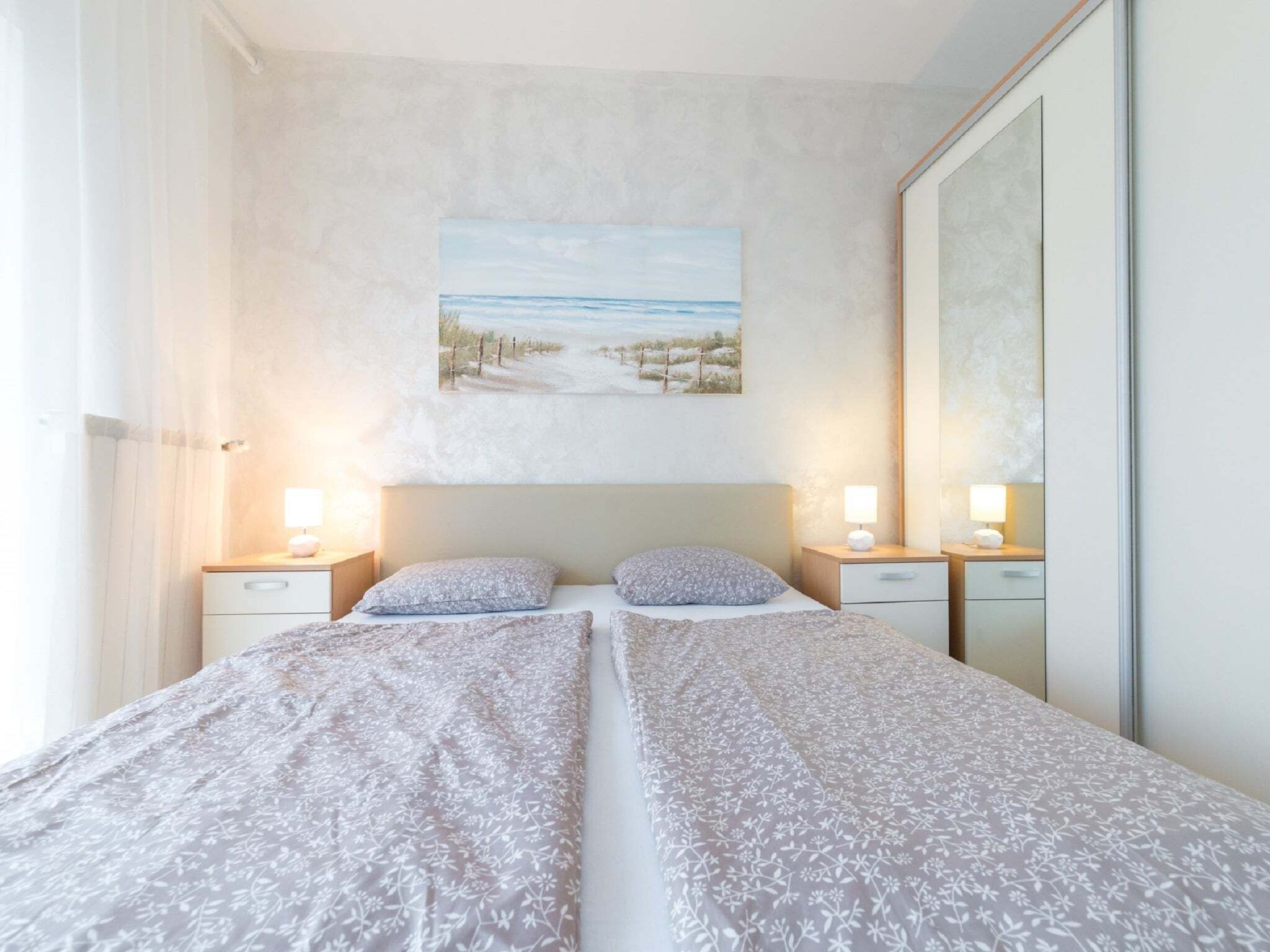Un lit double avec une literie à motifs floraux, deux tables de chevet avec des lampes, un grand miroir et un tableau représentant une scène de plage au mur.