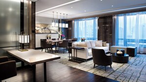 Una habitación de hotel moderna con zona de comedor, un conjunto de sofás y un amplio ventanal con vistas a la ciudad.