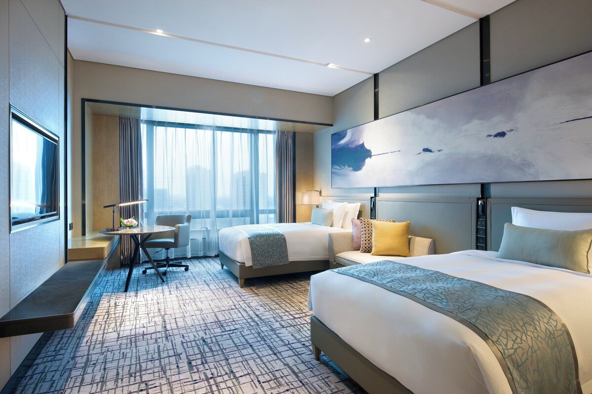 A hotel room with a large bed, a smaller bed, a desk, and a chair. There is a window with curtains and a painting on the wall.