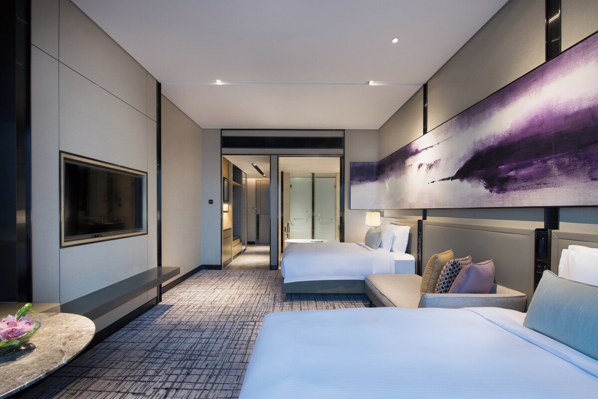 A modern hotel room with a large bed, a seating area, a television, and a painting on the wall.
