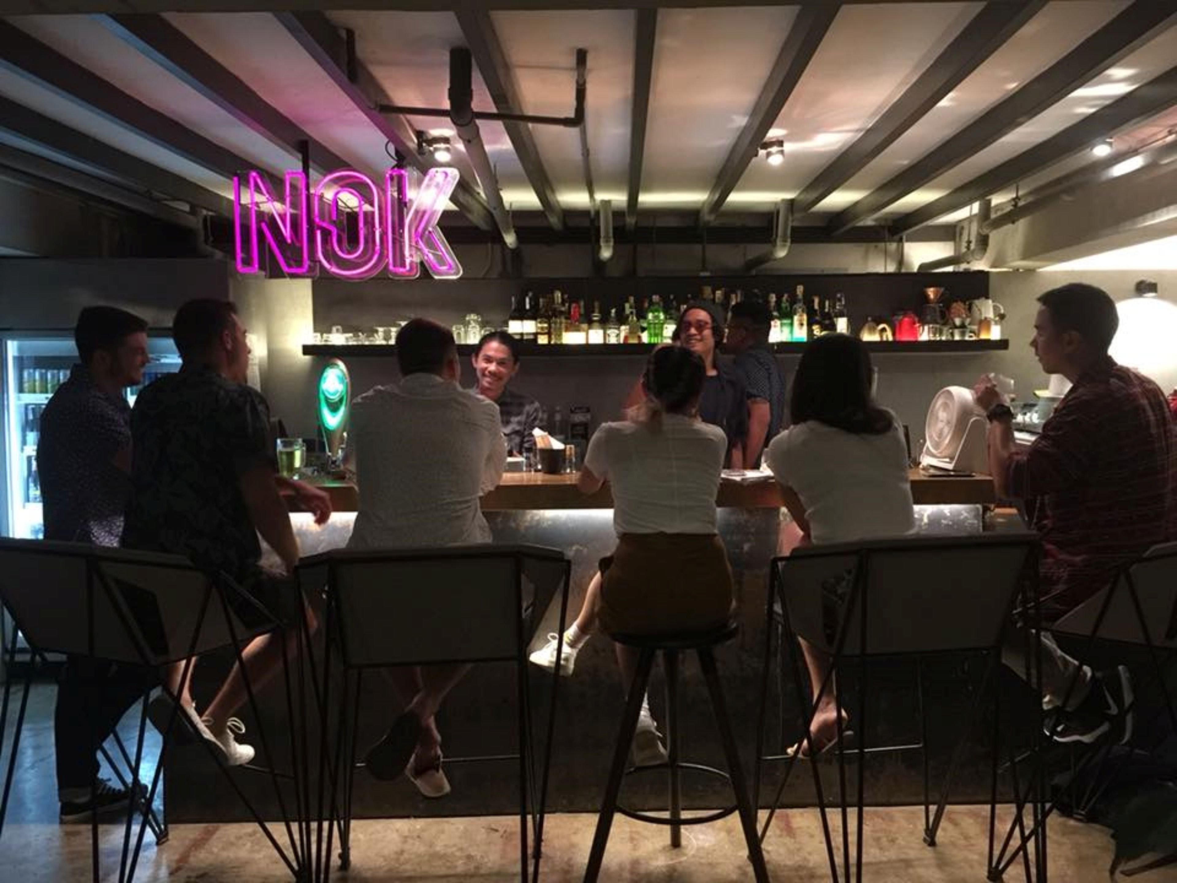 A bar area with a neon sign reading "NOK" and people seated at the bar.