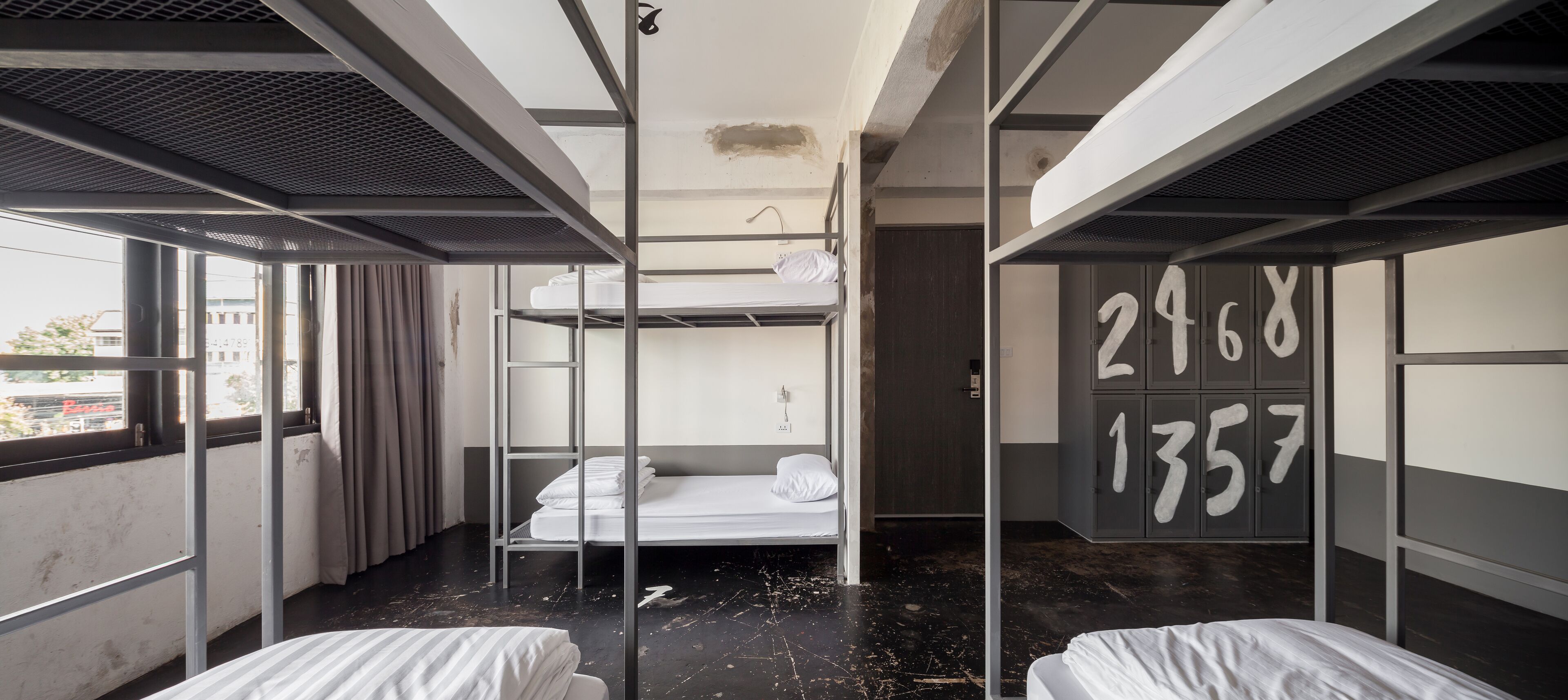 A dormitory room with bunk beds, a window with curtains, and a door with numbers on it.