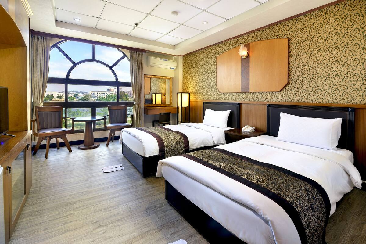 A hotel room with two beds, a desk, and a large window offering a city view.