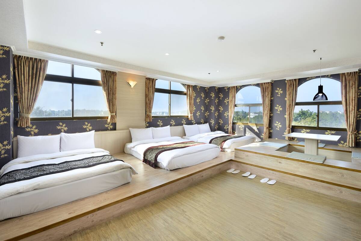 A spacious hotel room with two beds, a desk, and large windows offering a city view.