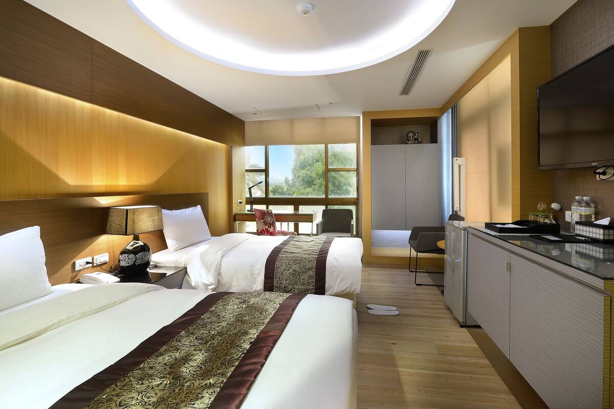 A hotel room with two beds, a kitchenette, a TV, and a large window with a view of trees.