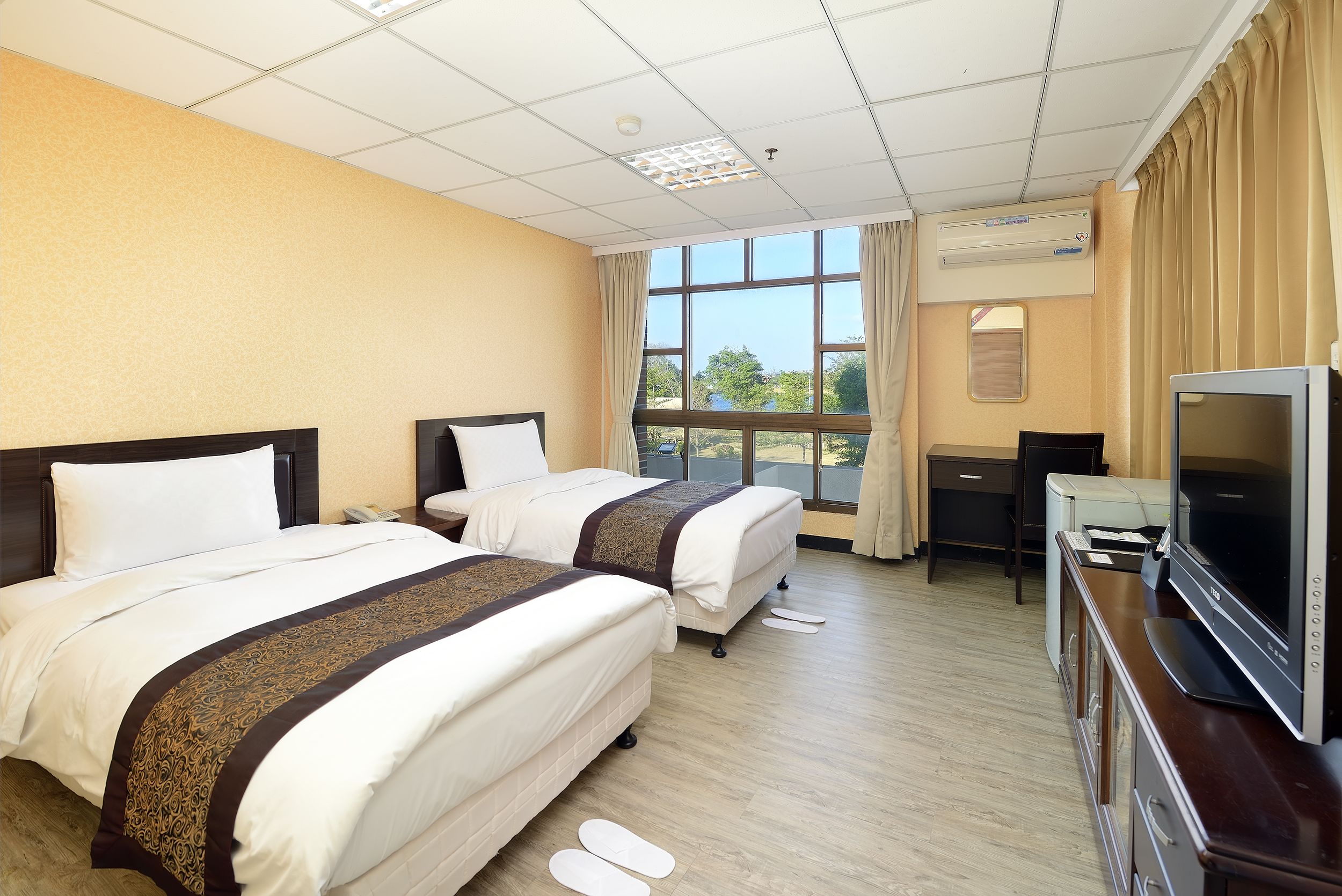 A hotel room with two beds, a desk, a television, and a window with a view of trees.