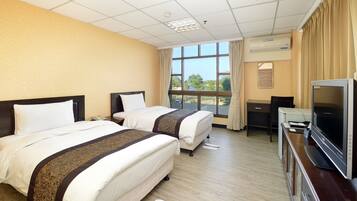 A hotel room with two beds, a desk, a television, and a window with a view of trees.