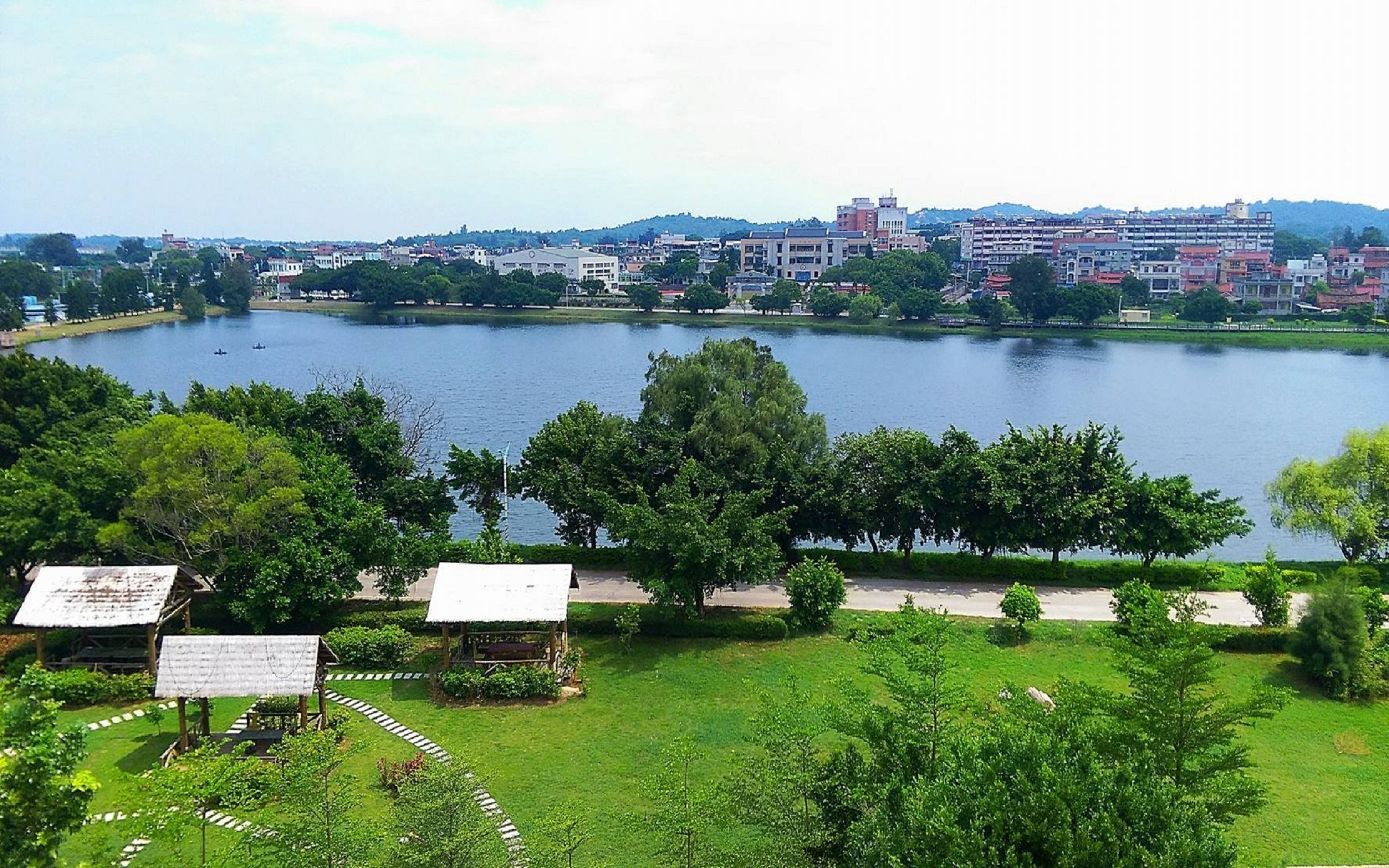 Une vue sur le lac, avec des bâtiments en arrière-plan, une zone herbeuse parsemée d’arbres et un petit pavillon.