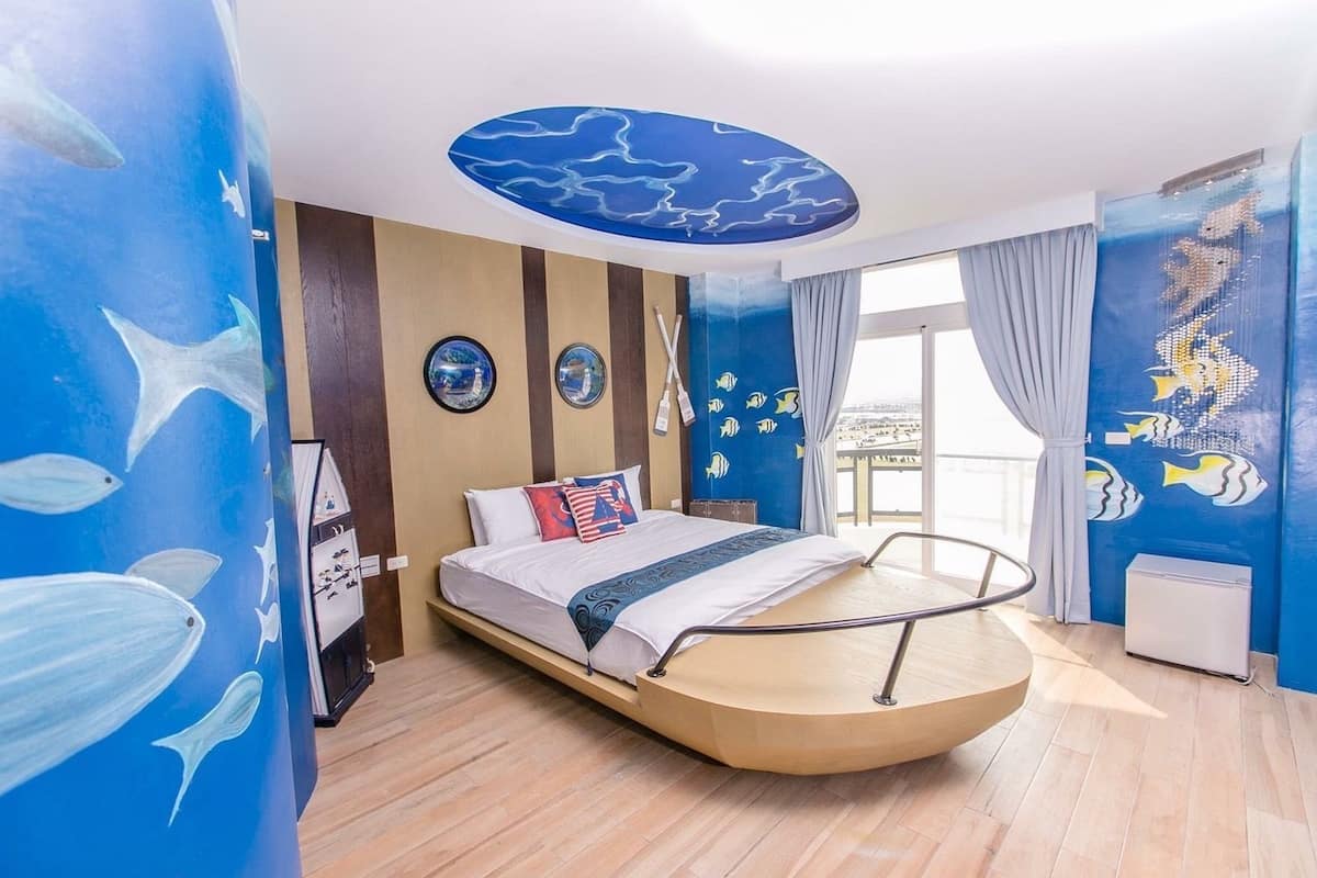 A room with a curved bed, a view of the sea through large windows, and a ceiling mural with fish.