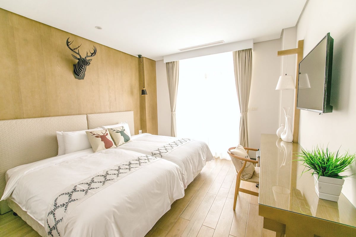 A hotel room with two beds, a wooden headboard, a mounted deer head, a TV, a glass table, and a potted plant.