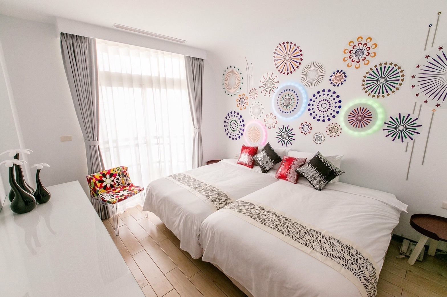 A hotel room with two beds, a desk, and a chair. The room has a large window with curtains, a wall decorated with circular patterns, and a floor made of light-colored wood.