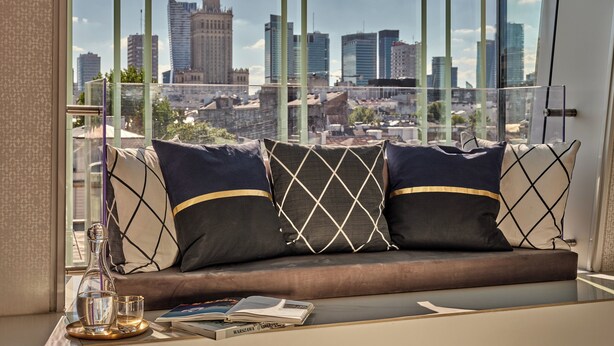 Un salon moderne avec vue sur la ville, un canapé agrémenté de coussins à motifs, une table basse en verre et un livre intitulé « Varsovie ».