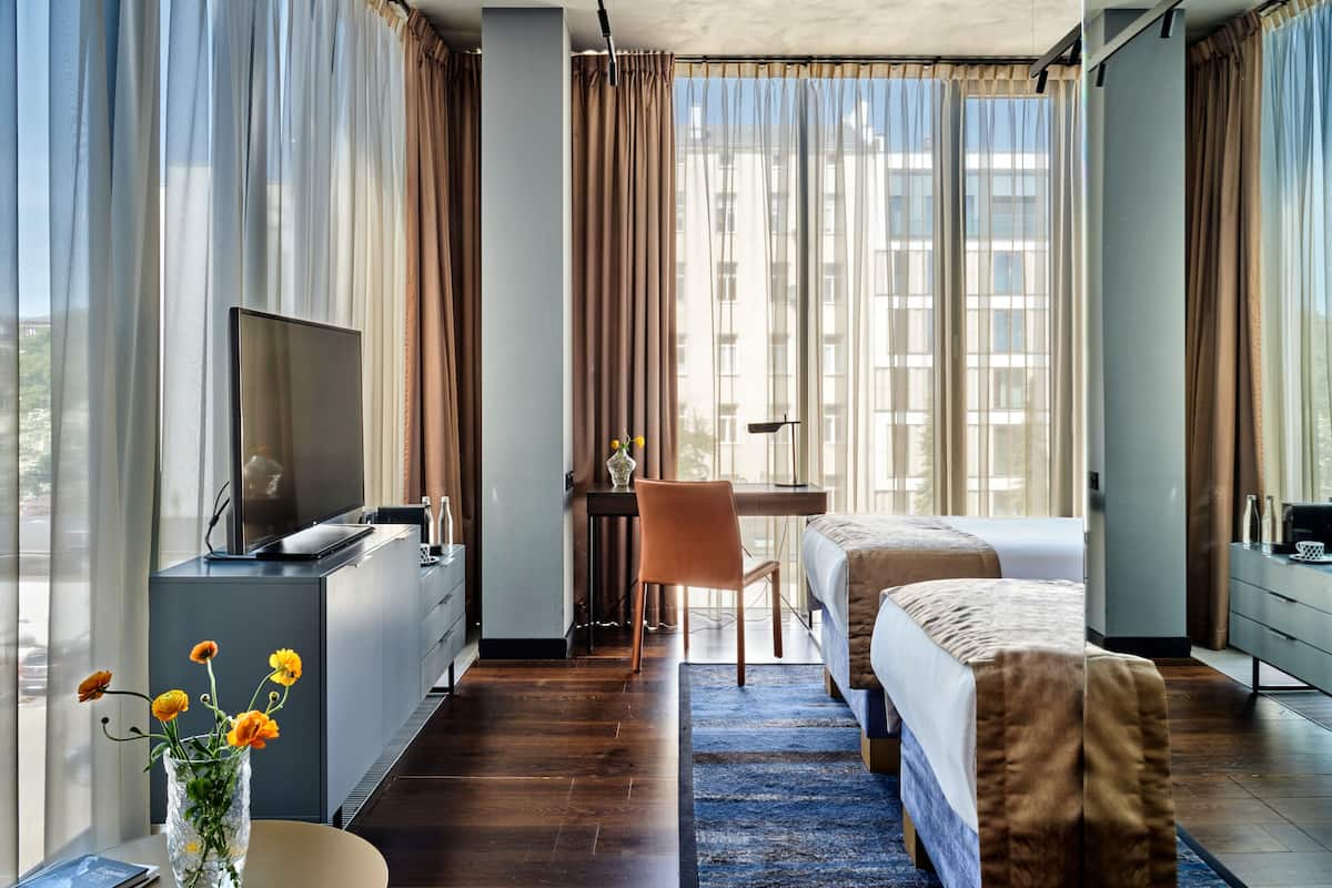 Een moderne hotelkamer met een bed, bureau, televisie en uitzicht op gebouwen door grote ramen.