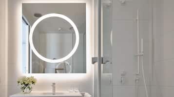 Casa de banho moderna com toucador branco, espelho redondo com luzes, cabine de duche em vidro e um vaso de flores na pia.