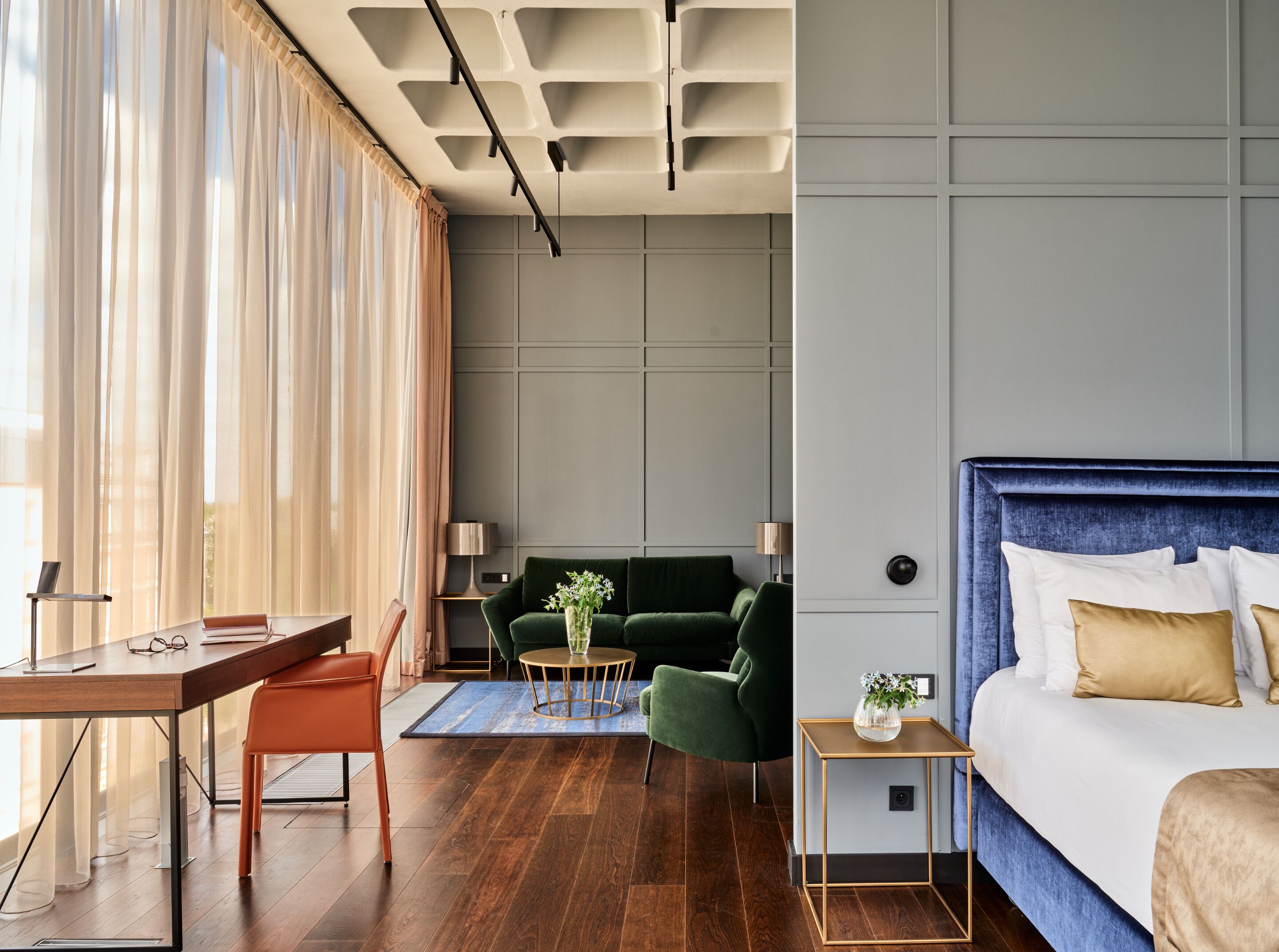 A modern hotel room with a wooden desk, a green sofa, a round coffee table, and a large bed with a blue headboard.