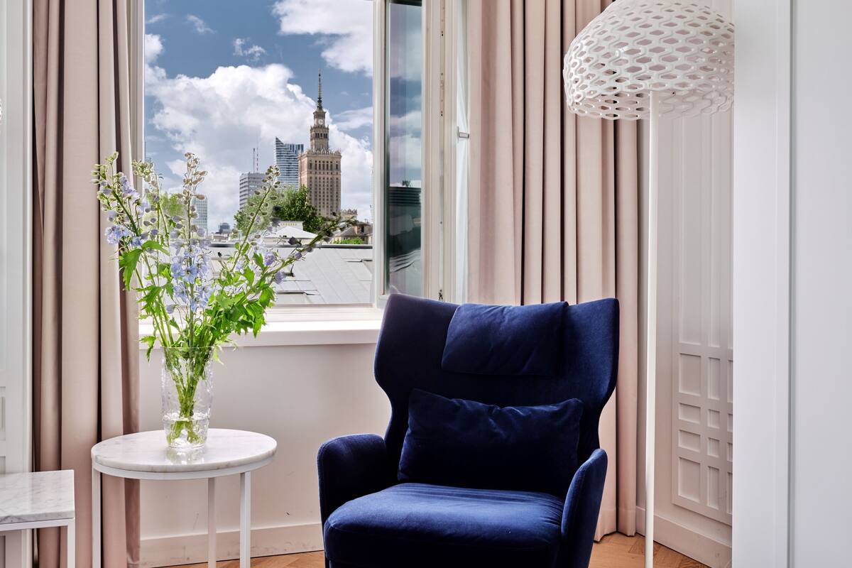 Een kamer met uitzicht op een stadsgezicht door een raam, een blauwe stoel, een witte tafel met een bloevas en een witte lamp.