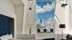 Une chambre d’hôtel moderne dotée d’un grand lit, d’un canapé, d’un fauteuil et offrant une vue sur le paysage urbain à travers une fenêtre.