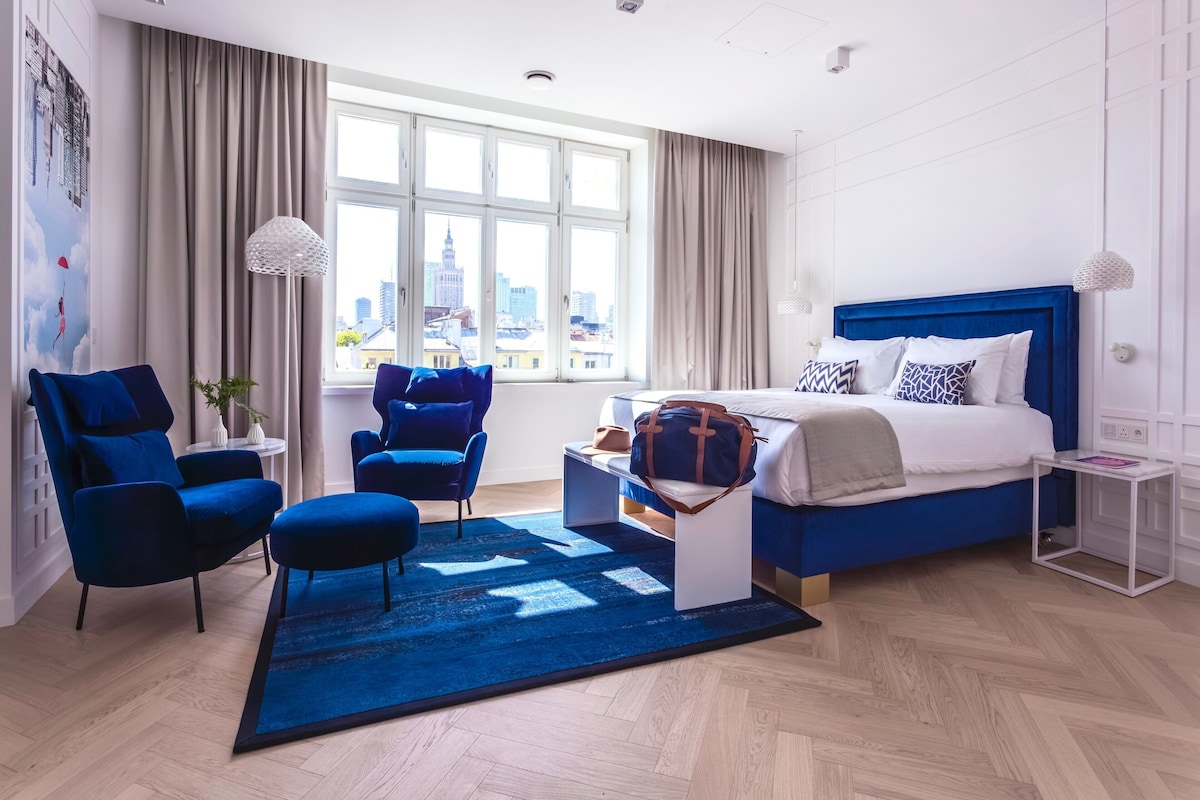 Ein modernes Hotelzimmer mit einem großen Bett, zwei blauen Sesseln, einem kleinen Tisch und Blick auf die Skyline durch große Fenster.