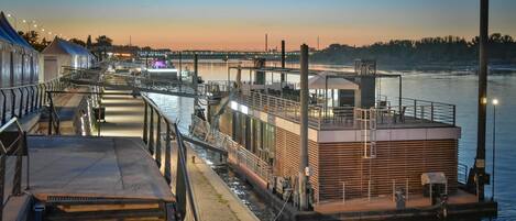Un muelle frente al agua con un barco, un pasillo cubierto y un edificio de techo plano.