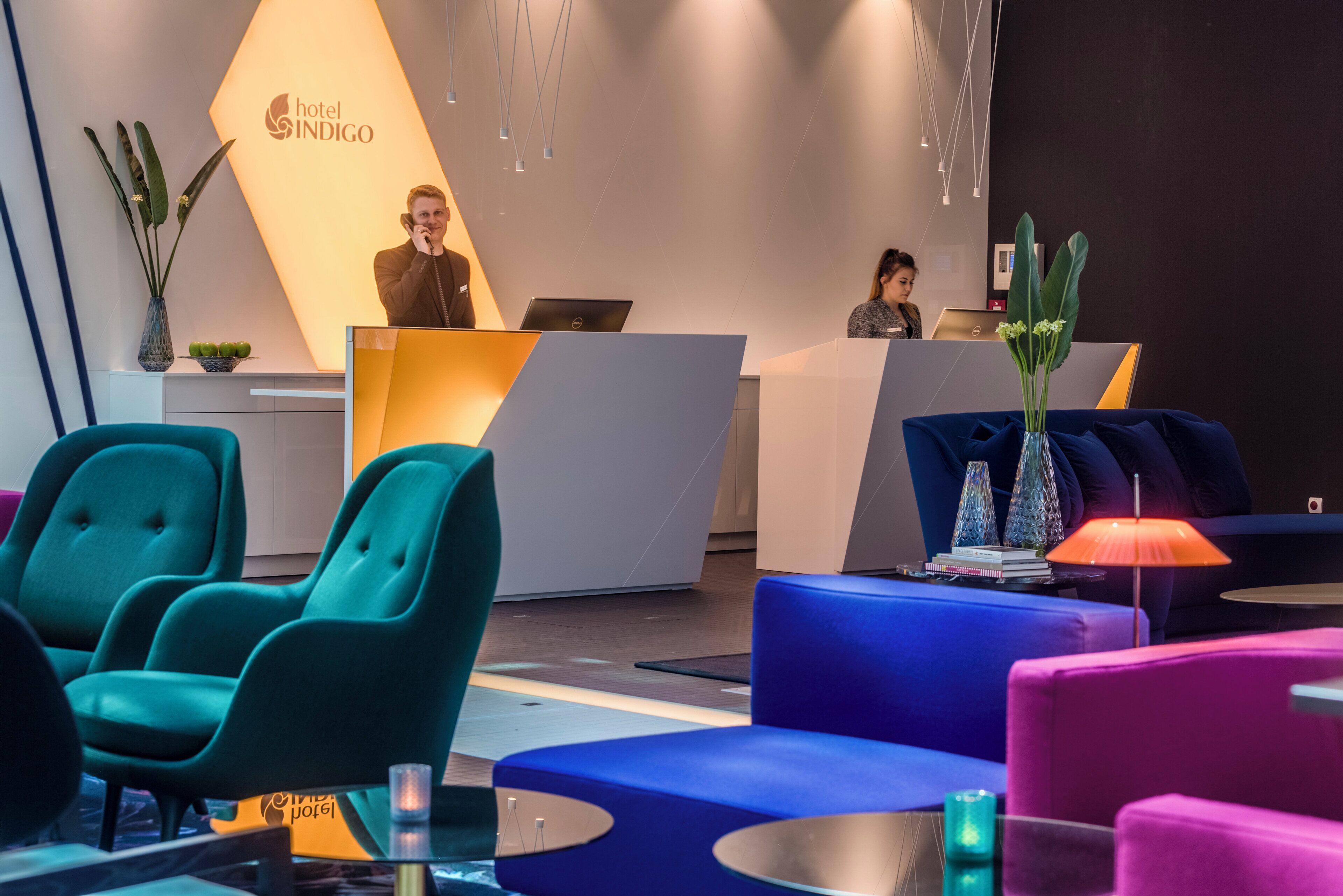 A hotel reception area with teal and blue seating, a reception desk, and a large illuminated logo.